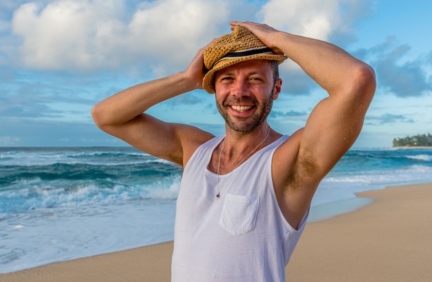 Giovane maschio caucasico in piedi sulla spiaggia del tramonto alle Hawaii che indossa un cappello di paglia