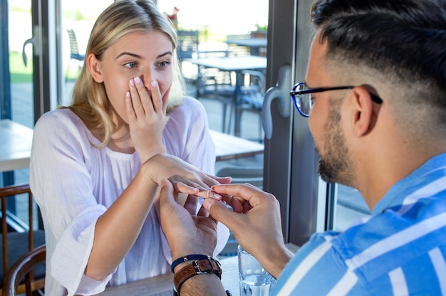 Giovane maschio che propone alla sua ragazza stupita in un ristorante con un anello di fidanzamento con diamante