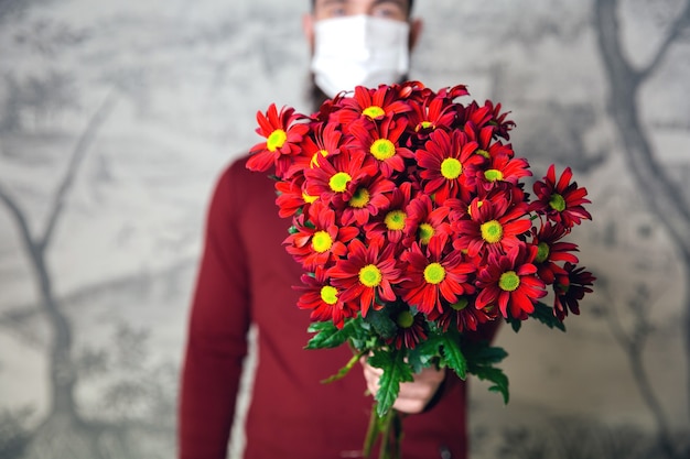 Giovane maschio con maschera protettiva per Coronavirus con bouquet di fiori, margherite rosse per romantico San Valentino o compleanno, presente e covid-19 concept romance