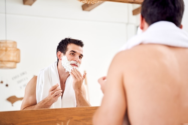 Giovane maschio con schiuma sulla barba vicino allo specchio al mattino