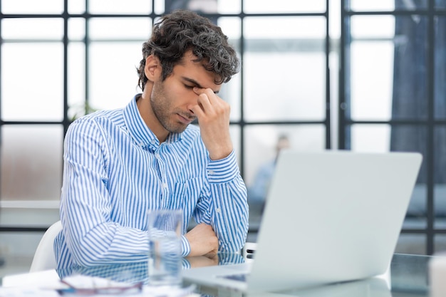 Giovane maschio frustrato infelice che tiene la testa a mano seduto con il computer dietro la scrivania in ufficio