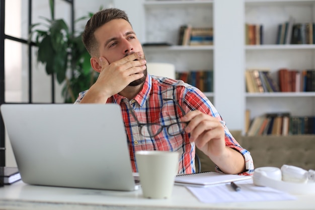 Giovane maschio frustrato infelice che tiene la testa con le mani seduto con il computer portatile dietro la scrivania a casa.