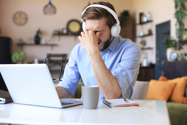 Giovane maschio frustrato infelice che tiene la testa con le mani seduto con il computer portatile dietro la scrivania a casa.