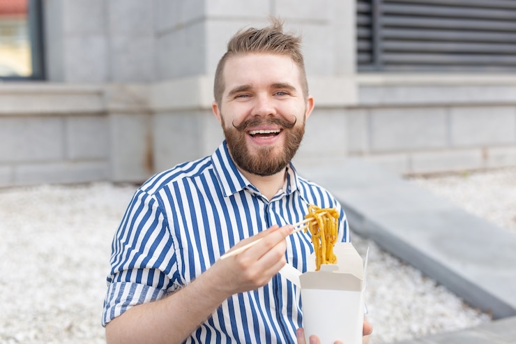 Foto giovane maschio hipster con baffi e barba sta mangiando spaghetti cinesi con le bacchette di legno da una scatola di pranzo bianca. concetto di cucina asiatica.