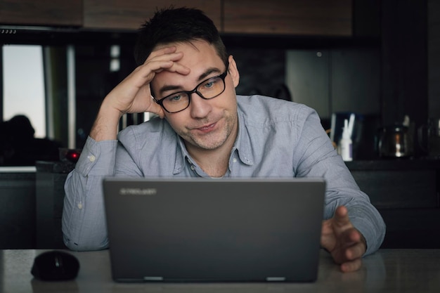 Giovane maschio in abiti casual concentrato ragazzo che aggiusta gli occhiali mentre lavora al computer portatile e guarda lo schermo del pc durante il lavoro a distanza dall'uomo che lavora troppo da casa sentendosi stanco primo piano