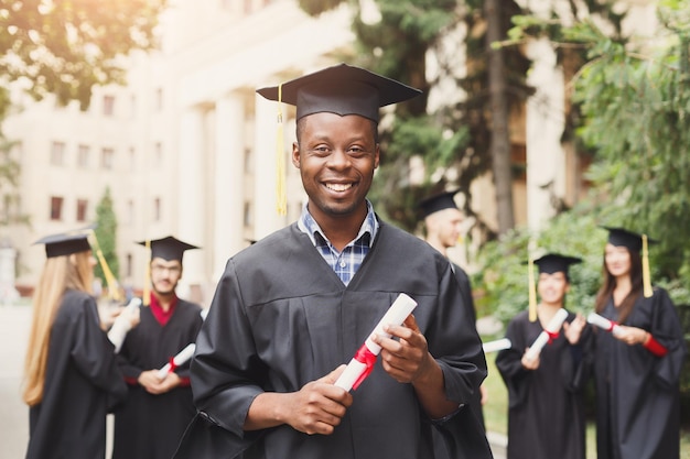 Giovane maschio nero sorridente il giorno della laurea all'università in piedi con un gruppo multietnico di amici. Istruzione, qualifica e concetto di abito.