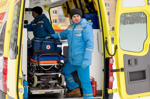 Giovane maschio paramedico con kit di pronto soccorso in piedi dalla barella nelle porte dell'ambulanza e guardando dritto