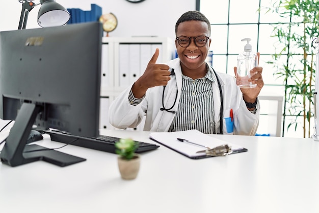 Giovane medico africano che tiene il gel disinfettante per le mani presso la clinica sorridendo felice e positivo, pollice in su facendo eccellente e segno di approvazione