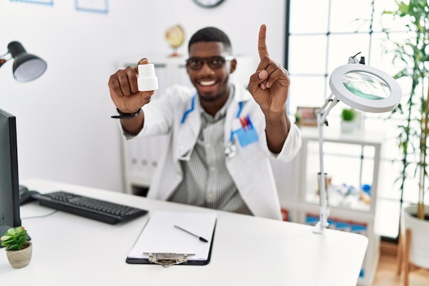 Giovane medico afroamericano che indossa l'uniforme del medico che tiene pillole da prescrizione presso la clinica sorpreso da un'idea o una domanda che punta il dito con la faccia felice, numero uno