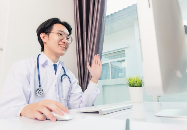 Giovane medico asiatico bello che indossa un abito da dottore e videoconferenza con stetoscopio o facetime con il computer al paziente che sorride seduto sulla scrivania presso l'ufficio dell'ospedale, assistenza medica sanitaria