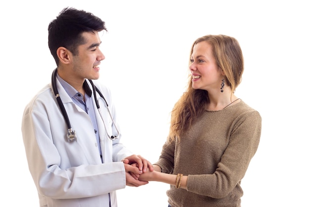 Giovane medico attraente con i capelli scuri in abito bianco con lo stetoscopio sul collo che parla con una giovane donna graziosa con lunghi capelli castani in maglione e jeans su sfondo bianco in studio