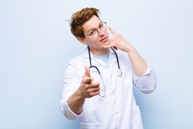 Giovane medico capo rosso che sorride allegramente e che indica la macchina fotografica mentre ti fa una chiamata in seguito gesto, parlando al telefono sulla parete blu