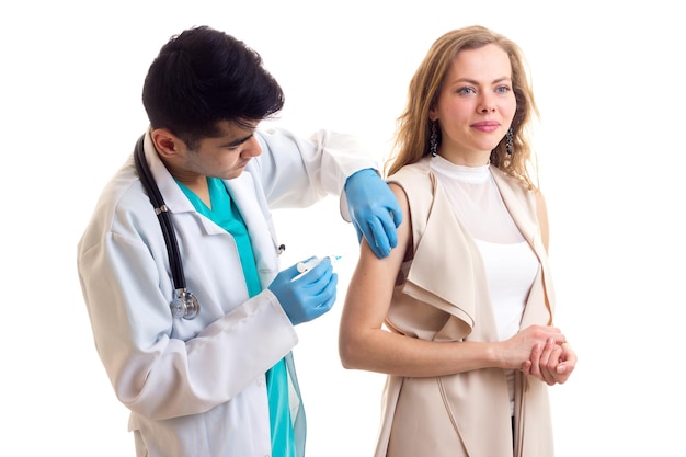 Giovane medico dall'aspetto elegante con capelli neri in abito bianco con stetoscopio e guanti blu che fa un'iniezione di giovane bella donna con lunghi capelli castani in camicetta beige su sfondo bianco in studio