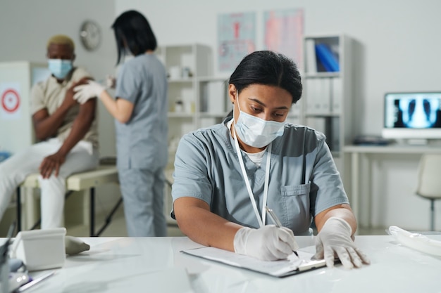 Giovane medico di razza mista in abbigliamento da lavoro protettivo seduto alla scrivania e compilando un documento mentre il suo collega vaccina un paziente maschio