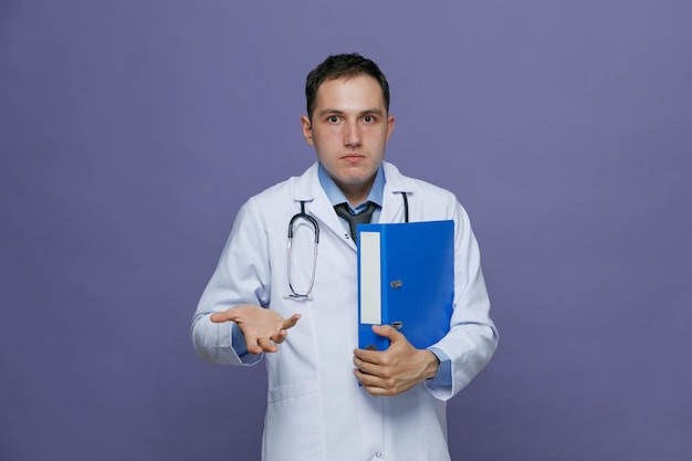 Giovane medico di sesso maschile senza indizi che indossa una veste medica e uno stetoscopio intorno al collo che tiene la cartella che lo abbraccia guardando la fotocamera che mostra la mano vuota isolata su sfondo viola