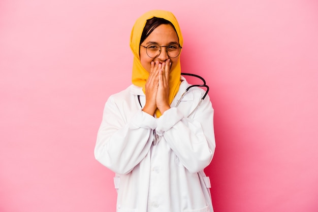 Giovane medico donna musulmana isolata su sfondo rosa che ride di qualcosa, coprendo la bocca con le mani.