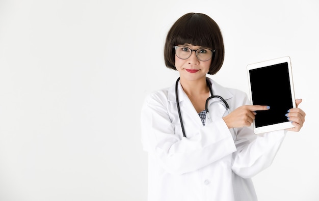 Giovane medico femminile asiatico positivo con lo stetoscopio che punta allo schermo nero vuoto del tablet moderno mentre presenta un'app medica su sfondo bianco