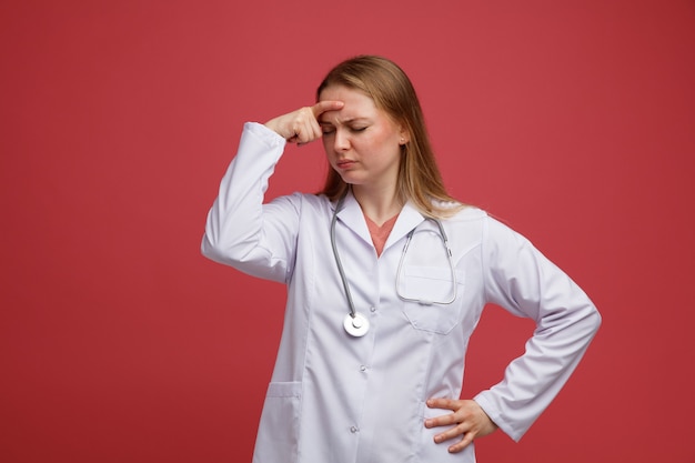 Giovane medico femminile biondo stanco che indossa veste medica e stetoscopio intorno al collo tenendo la mano sulla vita e sulla fronte con gli occhi chiusi