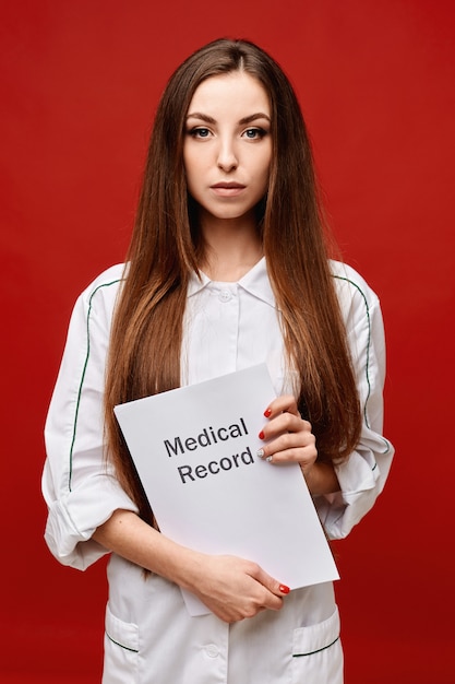 Giovane medico femminile in uniforme medica che tiene una cartella sanitaria in sue mani, spazio della copia, isolato. Concetto di assistenza sanitaria