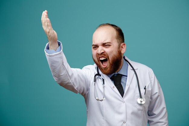 Giovane medico maschio arrabbiato che indossa cappotto medico e stetoscopio intorno al collo alzando la mano guardando il lato gridando isolato su sfondo blu