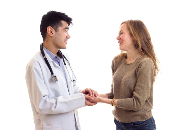 Giovane medico sorridente con i capelli scuri in abito bianco con lo stetoscopio sul collo che parla con una giovane donna graziosa con lunghi capelli castani in maglione e jeans su sfondo bianco in studio