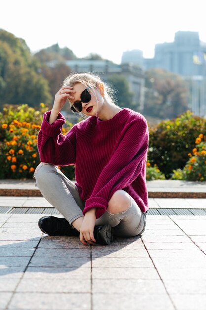 Giovane modella affascinante che indossa un maglione lavorato a maglia e jeans strappati seduto sul marciapiede