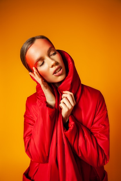 Giovane modella femminile in elegante cappotto rosso che tiene la mano in tasca e tocca il viso con un trucco creativo su sfondo giallo