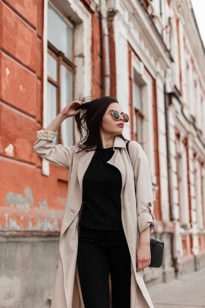 Giovane modello di bella donna abbastanza bella in occhiali da sole alla moda in trench beige alla moda cammina vicino a un edificio d'epoca in città. La ragazza adorabile gode di una passeggiata sulla strada. Abbigliamento moda primaverile per donna.