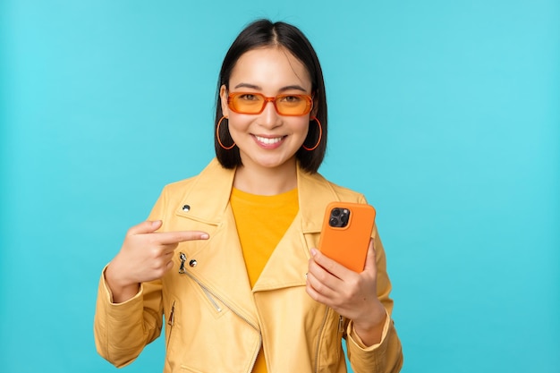 Giovane modello femminile asiatico alla moda in occhiali da sole alla moda vestito primaverile che mostra il telefono cellulare che punta allo smartphone e sorridente in piedi su sfondo blu