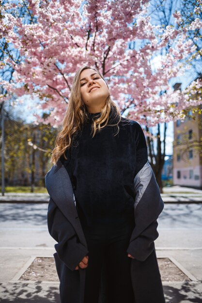 Giovane modello femminile snello con lunghi capelli ondulati e, vestito con un cappotto grigio, scarpe da ginnastica, che gira per strada. Primavera in fiore alberi donna ragazza ride e corre a gioire