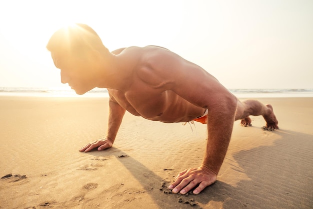 Giovane modello maschio muscoloso bello che fa l'allenamento che allunga il riscaldamento sulla spiaggia summer.sexy atleta addominali e corpo perfetto nel tramonto sulla riva del paradiso tropicale.