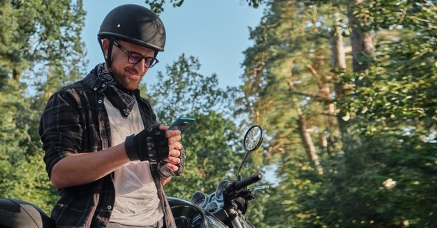 Giovane motociclista maschio in casco utilizzando il telefono cellulare e sorridente seduto su una moto