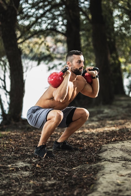 Giovane muscolare con il torso nudo che fa esercizi di squat con kettlebell nella natura, vicino al fiume.