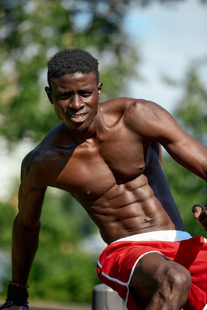 Giovane muscoloso che si esercita nel campo sportivo Uomo africano che guarda di lato mentre fa esercizi di barra orizzontale Modello maschile senza camicia che si esercita all'aperto