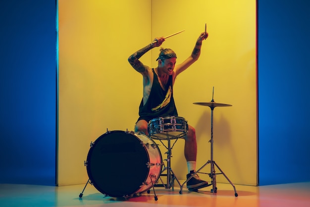 Giovane musicista con batteria che si esibisce su sfondo giallo alla luce al neon. Concetto di musica, hobby, festival, intrattenimento, emozioni. Batterista gioioso e ispirato. Ritratto colorato dell'artista.