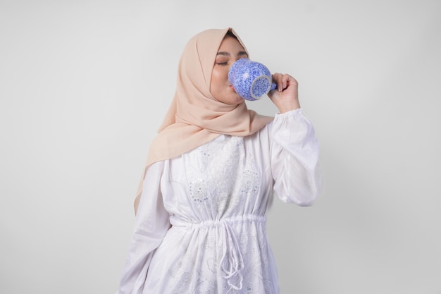 Giovane musulmana asiatica assetata in abito bianco e velo crema hijab che beve acqua minerale fresca da una tazza blu dopo il digiuno concetto di Ramadan