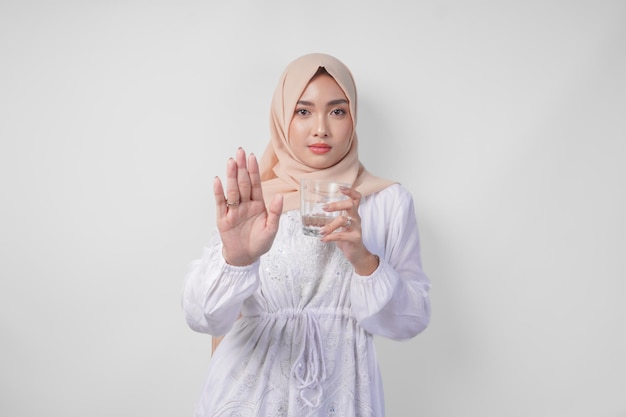 Giovane musulmana asiatica seria che indossa un vestito bianco e un hijab che mostra il palmo per fare un gesto di fermata mentre tiene in mano una tazza d'acqua in piedi su uno sfondo bianco isolato concetto di Ramadan