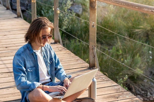 Giovane online che lavora come libero professionista con i capelli lunghi seduto sul pavimento che si sveglia sul suo laptop mentre è in vacanza