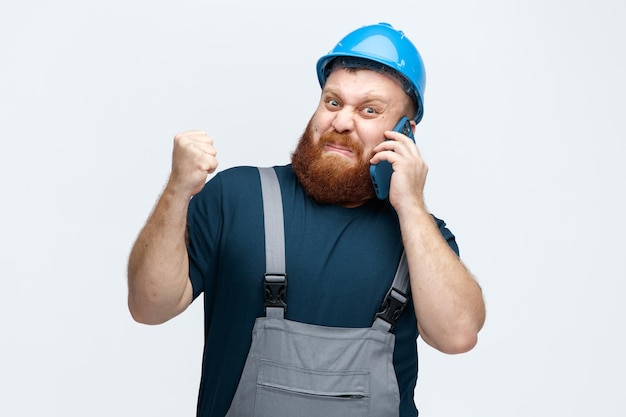 Giovane operaio edile maschio irritato che indossa casco di sicurezza e uniforme che parla al telefono guardando la fotocamera tenendo il pugno in aria isolato su sfondo bianco