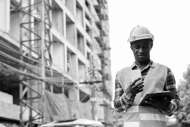 Giovane operaio edile uomo africano nero lettura negli appunti tenendo il telefono cellulare in cantiere