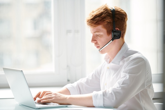 Giovane operatore del call center che comunica con i clienti online mentre è seduto davanti al computer portatile in ufficio