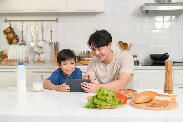 Giovane padre asiatico e suo figlio che usano la tavoletta digitale godendosi insieme in cucina a casa