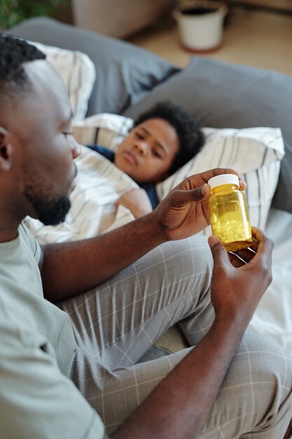 Giovane padre attento che tiene in mano una bottiglia di pillole mentre è seduto sul letto accanto al suo figlioletto malato con la febbre e gli darà un tablet