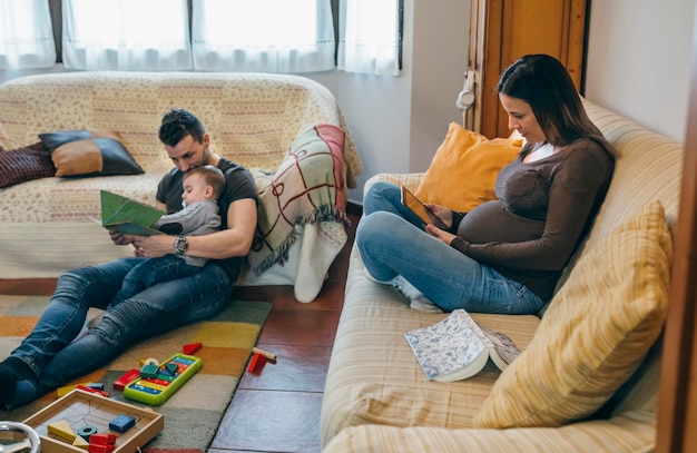 Giovane padre che legge un libro al suo piccolo figlio mentre la madre incinta sta guardando il tablet