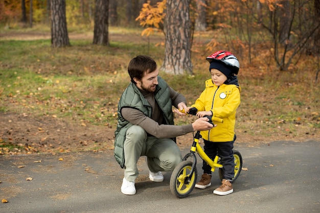 Giovane padre in abbigliamento casual che controlla la bici dell'equilibrio del suo simpatico figlioletto con il casco di sicurezza prima di guidarlo nel parco mentre entrambi sono in piedi sulla strada