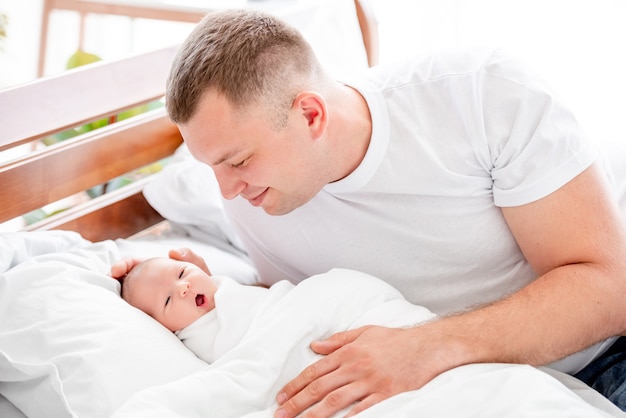 Giovane padre seduto nel letto e guardando con tenerezza la figlia appena nata. Papà genitore sorridente e bambina neonata a casa con la luce del giorno