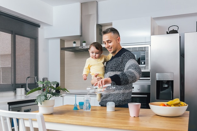 Giovane padre single che prepara un biberon mentre si tiene il suo bambino in braccio in una cucina moderna