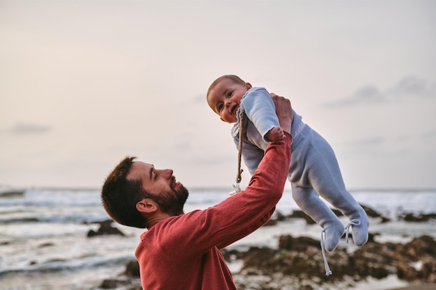 Giovane padre single che solleva il suo bambino e gioca con lui sulla spiaggia