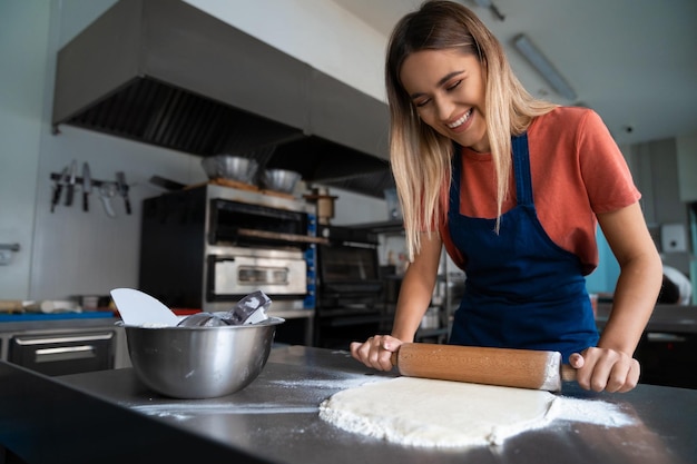 Giovane panettiera sorridente che impastano la pasta con un rotolo di legno in una cucina commerciale che fa il pane indossando un grembiule