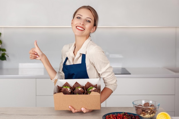 Giovane panettiere sorridente della donna in uniforme che tiene la scatola di imballaggio di consegna della carta con i cupcakes